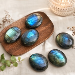 May include: Six polished oval labradorite stones featuring iridescent blue and green flashes. Three stones are arranged on a smooth, dark brown wooden tray, while the remaining three are positioned on a light-colored fabric surface nearby.
