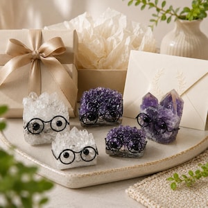 May include: Four crystal clusters in shades of clear and deep purple, each adorned with small black wire-rimmed glasses. The crystals are arranged on a tray alongside a beige gift box with a satin ribbon and a cream-colored envelope with a leaf design.