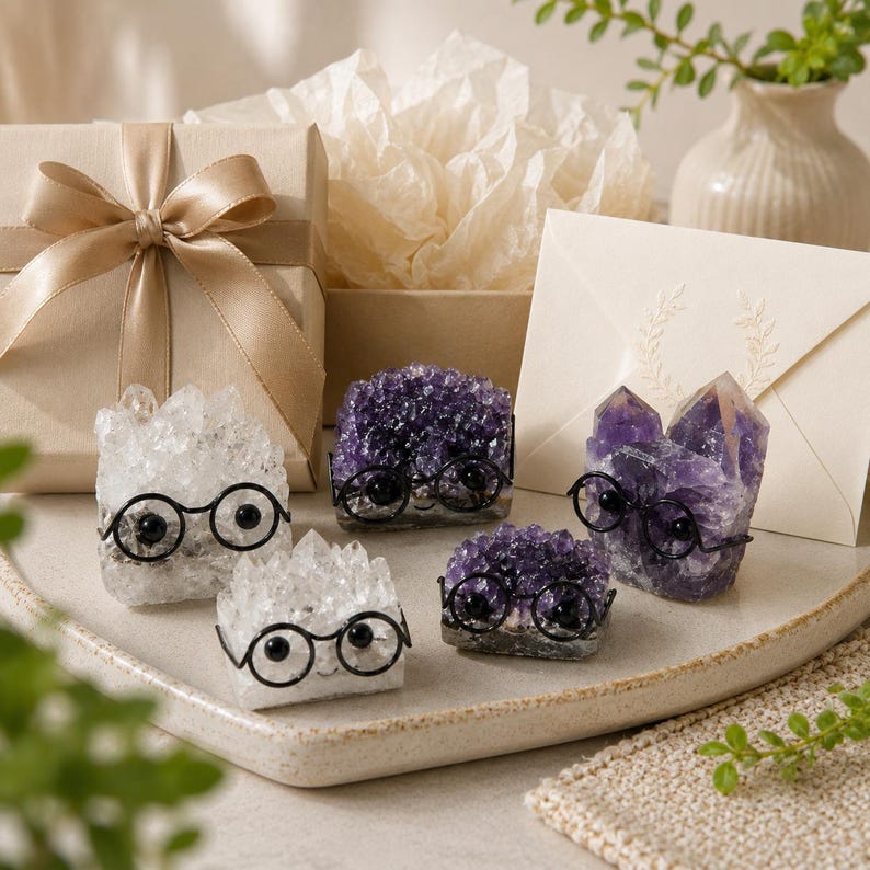 May include: Four crystal clusters in shades of clear and deep purple, each adorned with small black wire-rimmed glasses. The crystals are arranged on a tray alongside a beige gift box with a satin ribbon and a cream-colored envelope with a leaf design.