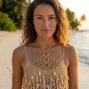 May include: A woman wearing a gold-colored necklace and top. The necklace features a series of connected circles and hangs over a top adorned with seashells. The background shows a beach with palm trees and the ocean.