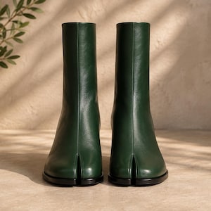 May include: A pair of dark forest green ankle boots featuring a distinct split-toe design. The boots have a smooth leather-like finish, a vertical seam running up the front center, and a solid black sole. The shaft of the boots reaches above the ankle.