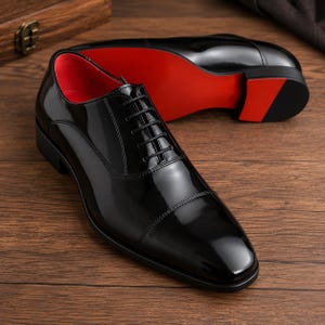 K&ouml;nnte beinhalten: Ein Paar gl&auml;nzende schwarze Lederschuhe mit roten Sohlen. Die Schuhe haben ein klassisches Oxford-Design mit Kappe und Schn&uuml;rverschluss. Ein Schuh ist im Fokus, w&auml;hrend der andere im Hintergrund leicht verschwommen ist, auf einer Holzoberfl&auml;che liegend.