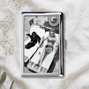 Puede incluir: Una pitillera rectangular de metal con una fotografía en blanco y negro de un chimpancé y una persona en bikini descansando en tumbonas a rayas. El estuche tiene un marco plateado pulido y un mecanismo de cierre lateral para abrirlo.