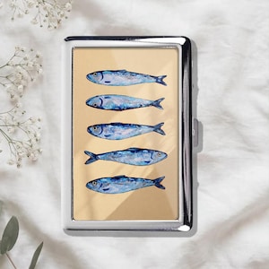 May include: A rectangular metal cigarette case featuring a polished silver frame and a central panel displaying a vertical arrangement of five blue fish illustrations painted in a watercolour style against a solid light tan background.