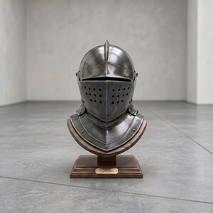 May include: A detailed, antique-style knight's helmet, crafted from dark metal, is displayed on a wooden bust. The helmet features a visor with multiple small holes and is mounted on a dark wooden base. A small, gold-colored plaque is affixed to the base.