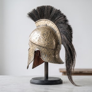 Puede incluir: Un casco de metal en tono bronce con textura martillada y una cresta alta de crin de caballo negra. El casco est&aacute; montado sobre un soporte de madera oscura con base circular, mostrando correas de cuero interiores y remaches met&aacute;licos.
