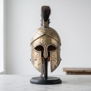 Puede incluir: Un casco de estilo corintio en tono bronce met&aacute;lico con una textura martillada y desgastada, un protector nasal central y un penacho alto de crin de caballo negra. El casco est&aacute; montado sobre un soporte circular negro para su exhibici&oacute;n.