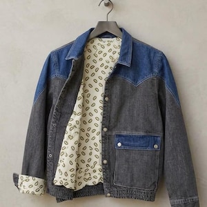 Puede incluir: Chaqueta vaquera de dos tonos. Los hombros y el cuello son de color azul claro, contrastando con el cuerpo gris oscuro. El forro interior de la chaqueta es de color crema con un patrón repetido. Tiene dos bolsillos delanteros y cierre de botones.