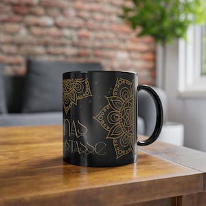 Könnte beinhalten: Schwarze Keramiktasse mit goldenem Mandala-Design und dem Text "Lunas Tasse". Die Tasse hat einen schwarzen Henkel und ein goldenes Blumenmuster. Die Tasse steht auf einer Holzoberfläche.