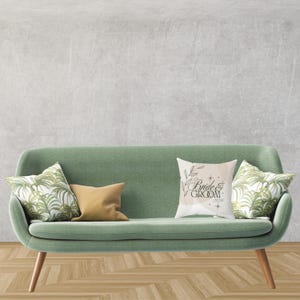Könnte beinhalten: Ein salbeigrünes Sofa mit Holzbeinen, verziert mit dekorativen Kissen. Ein Kissen trägt den Text "Bride & Groom 2026" in schwarzer Schrift. Andere Kissen zeigen botanische und unifarbene Designs. Das Sofa steht vor einer grauen Wand.