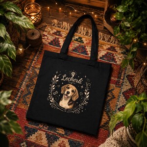 Könnte beinhalten: Eine schwarze Tragetasche mit einer zentralen Grafik eines Beagle-Hundes, umgeben von einem dekorativen Kranz aus Blättern, Sternen und Herzen. Der Text "Leckerli TASCHE" ist über dem Hund gedruckt, mit kleinerem Text unter der Illustration.
