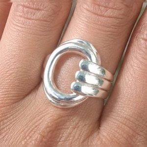 Puede incluir: Un anillo de plata pulida que presenta un diseño grande y redondeado de bucle abierto que se conecta a un vástago de triple banda. La superficie metálica es lisa y reflectante, creando una estructura continua e interconectada.
