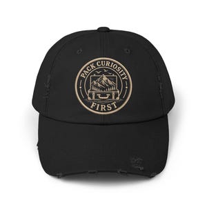 May include: A black distressed baseball cap featuring a circular embroidered patch on the front. The patch displays a suitcase containing a mountain range and pine trees, with the text PACK CURIOSITY FIRST arched around the design.