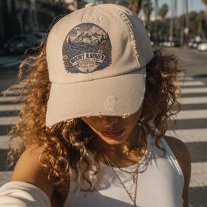 May include: A beige baseball cap with a distressed brim and a circular graphic on the front. The graphic features a mountain landscape, flowers, and the text "MOUNT RAINIER NATIONAL PARK WANDER MORE TRAVEL CLUB". The cap has a structured crown.