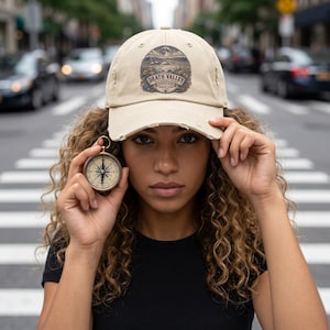 May include: A beige baseball cap with a distressed brim and a circular graphic on the front featuring a desert landscape, a moon, and the text Death Valley National Park Travel Club. A small round brass compass is held up next to the cap.