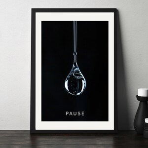 May include: A framed art print featuring a large, clear water droplet suspended against a black background. The word "PAUSE" is printed in white at the bottom. Two black decorative vases with white tops are on a dark wooden surface.