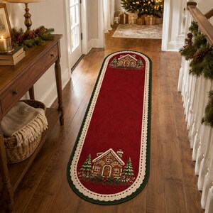 Puede incluir: Una alfombra de pasillo ovalada con un centro de color rojo intenso que presenta una ilustración de una casa de jengibre en cada extremo, flanqueada por pinos verdes. La alfombra tiene un borde festoneado color crema y un ribete verde oscuro.