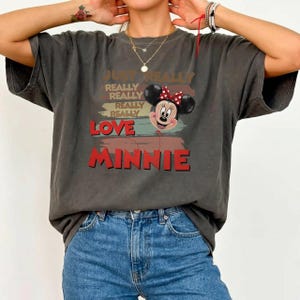 May include: A charcoal gray short-sleeve t-shirt featuring a graphic of Minnie Mouse with a red polka dot bow. The text reads JUST REALLY REALLY REALLY REALLY LOVE MINNIE in stacked red and tan lettering over a distressed background design.
