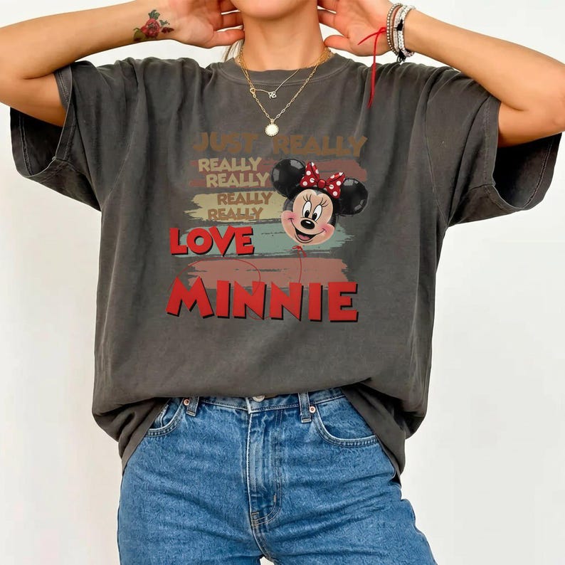 May include: A charcoal gray short-sleeve t-shirt featuring a graphic of Minnie Mouse with a red polka dot bow. The text reads JUST REALLY REALLY REALLY REALLY LOVE MINNIE in stacked red and tan lettering over a distressed background design.