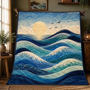 Op de afbeelding: Een doorgestikte deken met een oceaanlandschap met golvende blauwe en turquoise golven, een grote lichtgele zon en verschillende zwarte vogels die in een lichtblauwe lucht vliegen. De stof heeft een ruitvormig stikpatroon over het geheel.