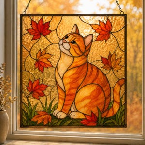 May include: A square stained glass suncatcher featuring an orange tabby cat sitting among fallen red and orange maple leaves. The cat is depicted in profile, looking upward, with a textured amber background and a dark metal frame with a hanging chain.