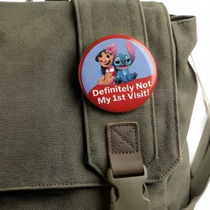 K&ouml;nnte beinhalten: Ein runder Ansteckbutton mit einer Cartoon-Illustration von Lilo und Stitch vor einem blau-roten Hintergrund. Der Text "Definitely Not My 1st Visit!" ist in Wei&szlig; auf dem unteren roten Bereich des Button-Designs aufgedruckt.