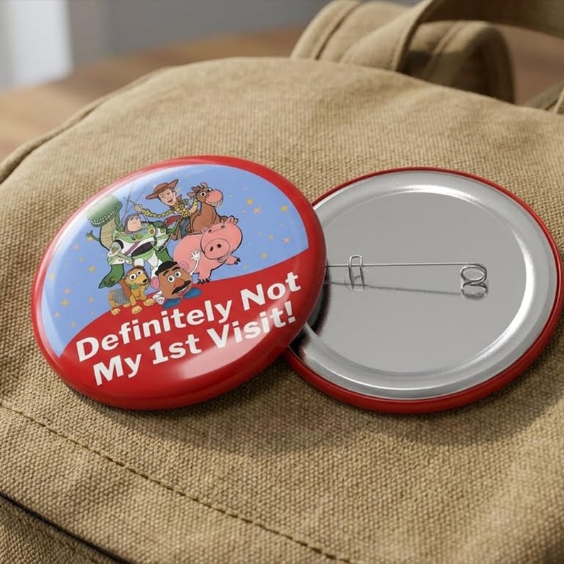 May include: A round pinback button featuring a blue background with a group of animated characters including a space ranger, a cowboy, and a pig. The lower portion is red with the white text "Definitely Not My 1st Visit!" and a metal pin back.