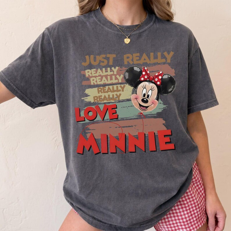 May include: A charcoal gray short-sleeve t-shirt featuring a graphic of Minnie Mouse as a balloon. The text reads JUST REALLY REALLY REALLY REALLY LOVE MINNIE in a mix of tan, red, and black colors over horizontal brushstroke background shapes.