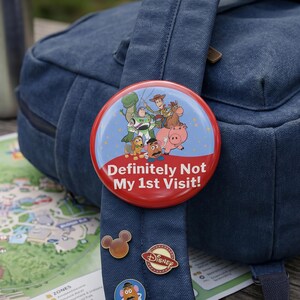 May include: A round button featuring Toy Story characters including Woody, Buzz Lightyear, Rex, Slinky Dog, Mr. Potato Head, Bullseye, and Hamm. The button has a red bottom section with the white text "Definitely Not My 1st Visit!" printed on it.