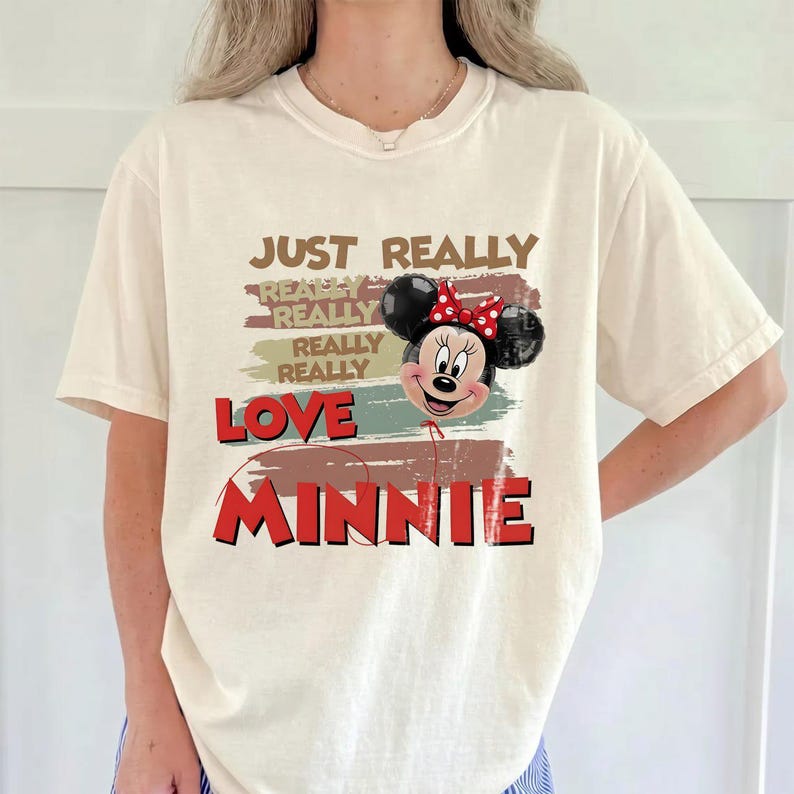 May include: A cream-colored t-shirt featuring a graphic print with the text JUST REALLY REALLY REALLY REALLY LOVE MINNIE. The design includes a balloon shaped like Minnie Mouse with a red polka dot bow and a red string extending downward.