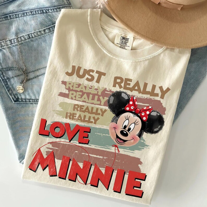 May include: A cream-colored t-shirt featuring the text "JUST REALLY REALLY REALLY REALLY REALLY LOVE MINNIE" in brown and red. A graphic of a Minnie Mouse balloon with a red and white polka dot bow is positioned next to the word "LOVE".
