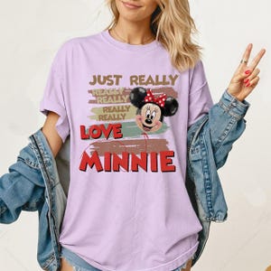 May include: A light purple short-sleeve t-shirt featuring a graphic of Minnie Mouse as a balloon. The text reads JUST REALLY REALLY REALLY REALLY LOVE MINNIE. The design includes horizontal brushstroke accents in muted earth tones behind the character.