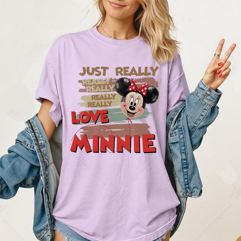 May include: A light purple short-sleeve t-shirt featuring a graphic of Minnie Mouse as a balloon. The text reads JUST REALLY REALLY REALLY REALLY LOVE MINNIE. The design includes horizontal brushstroke accents in muted earth tones behind the character.