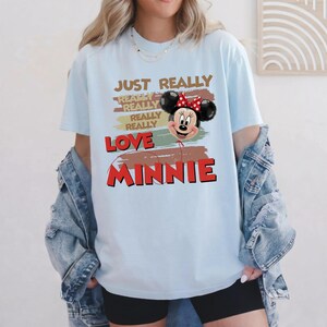 May include: A light blue t-shirt featuring a graphic of a Minnie Mouse balloon with a red polka dot bow. The text reads JUST REALLY REALLY REALLY REALLY LOVE MINNIE, layered over horizontal brush strokes in shades of brown, green, and red.