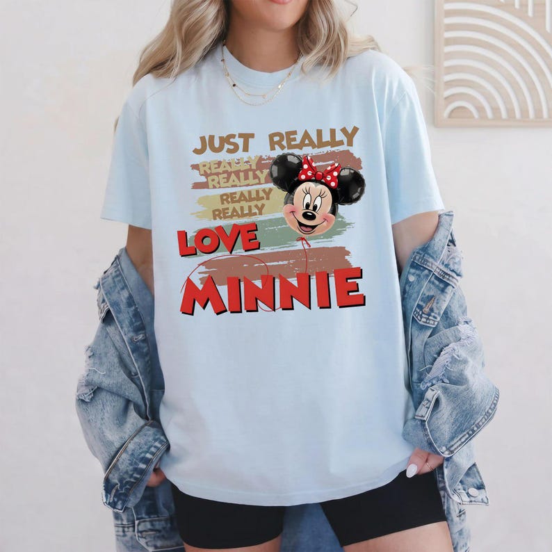 May include: A light blue t-shirt featuring a graphic of a Minnie Mouse balloon with a red polka dot bow. The text reads JUST REALLY REALLY REALLY REALLY LOVE MINNIE, layered over horizontal brush strokes in shades of brown, green, and red.