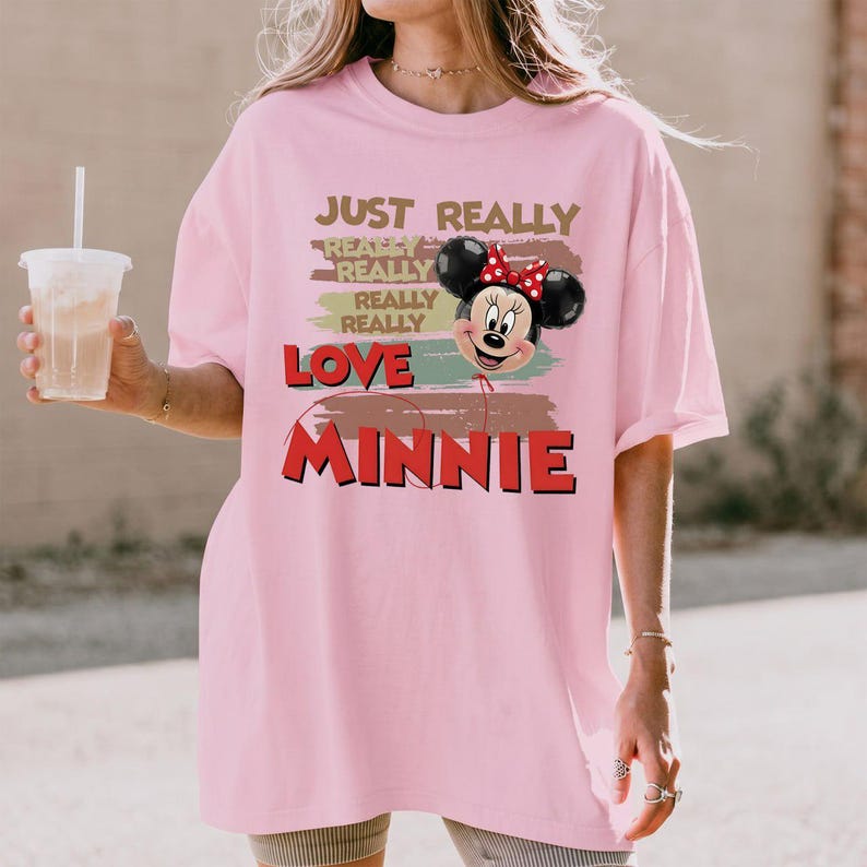 May include: A light pink t-shirt featuring a graphic of Minnie Mouse as a balloon. The text reads JUST REALLY REALLY REALLY REALLY LOVE MINNIE. The design includes horizontal brushstroke accents in muted green, brown, and red colors.