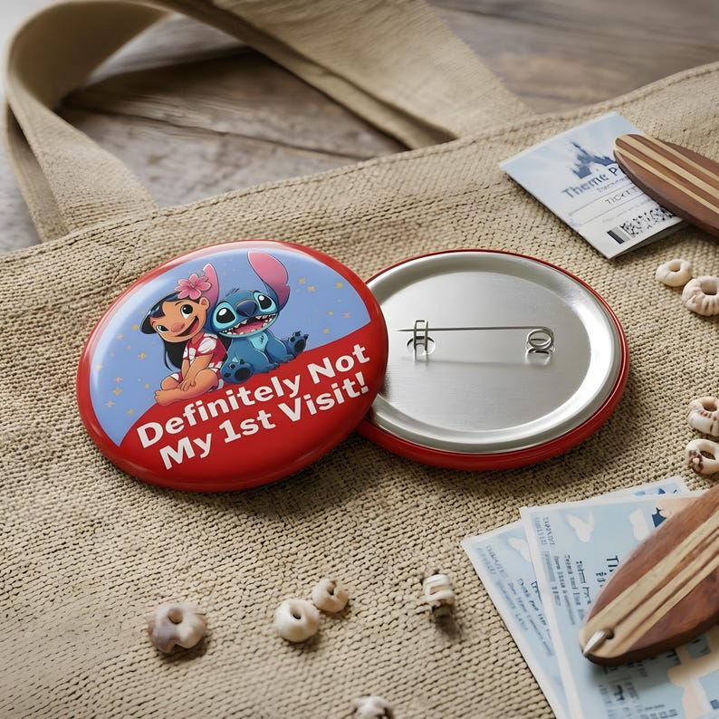 K&ouml;nnte beinhalten: Ein runder Ansteckbutton mit einem blau-roten Design und einer Illustration von Lilo und Stitch. Der Text "Definitely Not My 1st Visit!" ist in Wei&szlig; auf dem roten Bereich gedruckt. Die R&uuml;ckseite besteht aus silbernem Metall mit einer Nadel.