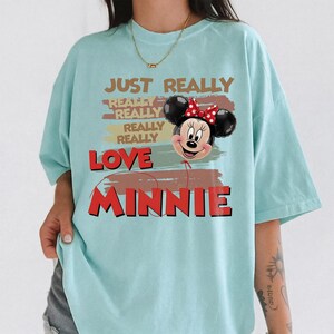 May include: A light blue t-shirt featuring a graphic of a Minnie Mouse balloon with a red polka dot bow. The text reads: JUST REALLY REALLY REALLY REALLY REALLY LOVE MINNIE. The design includes brown brushstroke accents behind the text and balloon.