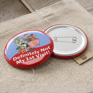 May include: A round pinback button featuring a blue and red design with Toy Story characters including Woody, Buzz Lightyear, Rex, Slinky Dog, Hamm, and Mr. Potato Head. The text "Definitely Not My 1st Visit!" is printed in white on the red section.