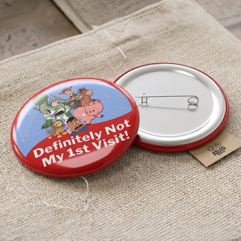 May include: A round pinback button featuring a blue and red design with Toy Story characters including Woody, Buzz Lightyear, Rex, Slinky Dog, Hamm, and Mr. Potato Head. The text "Definitely Not My 1st Visit!" is printed in white on the red section.
