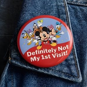 May include: A round pinback button featuring a blue and red background with illustrations of Mickey Mouse, Minnie Mouse, Donald Duck, Daisy Duck, Goofy, and Pluto. The text "Definitely Not My 1st Visit!" is printed in white on the red lower section.