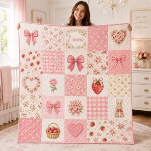 May include: A quilted blanket featuring a grid of pink and white squares with various patterns including bows, strawberries, floral arrangements, a heart, and a rabbit. The center square displays the name Emma surrounded by a floral wreath.