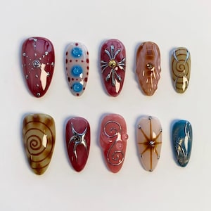Pode incluir: Um conjunto de dez unhas artificiais com vários designs. As unhas apresentam cores como vermelho, rosa, castanho e azul, com elementos decorativos como espirais prateadas, pontos e estrelas. Cada unha tem um acabamento brilhante.