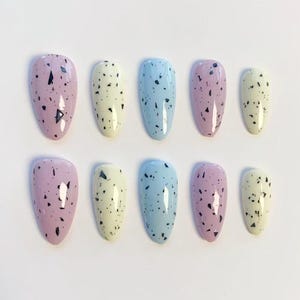 Puede incluir: Un conjunto de diez uñas postizas ovaladas en varios colores pastel: lavanda, amarillo pálido y azul claro. Cada uña está moteada con pequeñas motas negras. Las uñas están dispuestas en dos filas de cinco.