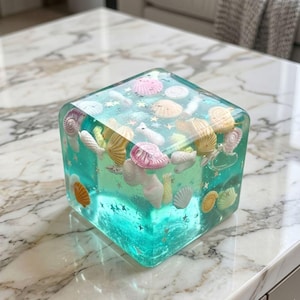 Op de afbeelding: Een heldere, kubusvormige presse-papier van hars met een turquoise tint, met diverse schelpen in roze, geel en wit. De kubus is versierd met kleine, goudkleurige stervormige glitter.