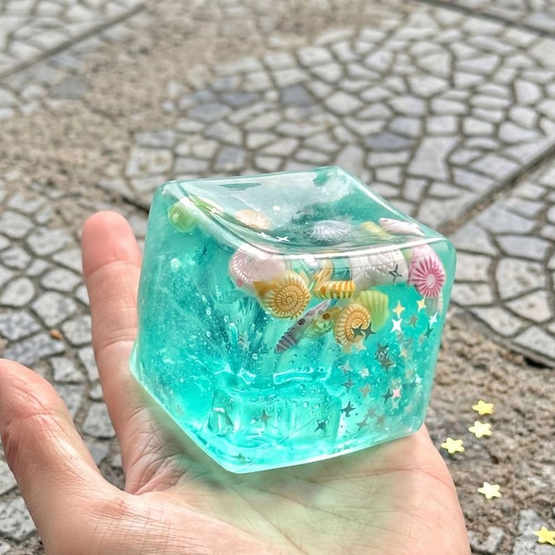 May include: A clear, cube-shaped resin paperweight with a turquoise interior, encasing various seashells and gold star-shaped glitter. The paperweight is held in a hand, showcasing the embedded marine life.