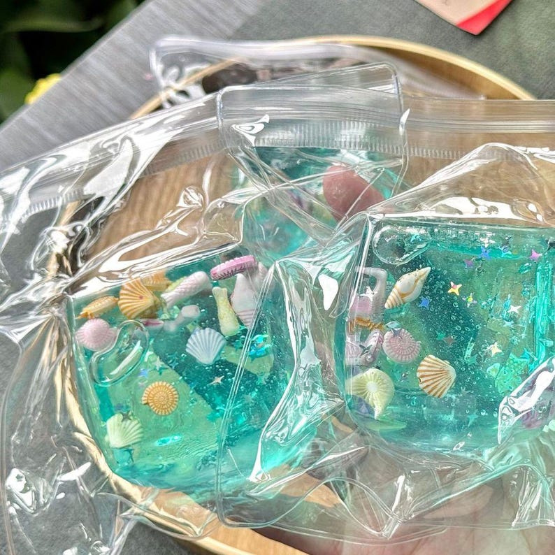 May include: Clear plastic pouches filled with turquoise-colored gel, miniature seashells, and glitter. The pouches are sealed and appear to be part of a craft or decorative project, possibly for resin art or jewelry making.