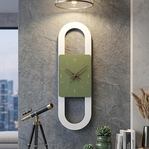 Op de afbeelding: Een wandklok met een verticaal wit ovaal frame rondom een vierkante olijfgroene wijzerplaat. De wijzerplaat is voorzien van goudkleurige uurmarkeringen, het cijfer 12 bovenaan en donkerbruine wijzers. Het ontwerp is strak en geometrisch.