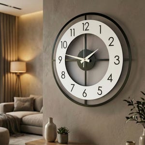 Könnte beinhalten: Eine runde Wanduhr mit einem äußeren Rahmen aus schwarzem Metall und einem weißen Zifferblatt mit schwarzen arabischen Ziffern von 1 bis 12. Die Uhr hat eine schwarze zentrale Nabe mit dünnen schwarzen Querstreben und weißen Zeigern.
