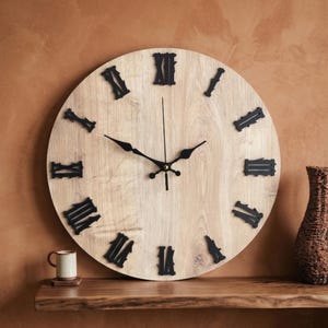 May include: A large, circular wall clock with a light wood grain face and black Roman numerals. The clock hands are black, and the clock is mounted on a brown wall. A small cup and a decorative vase are on a wooden shelf below the clock.